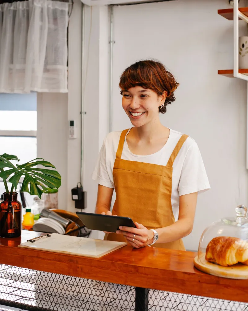 The same bakery owner smiling at the counter, tablet in hand, after approving